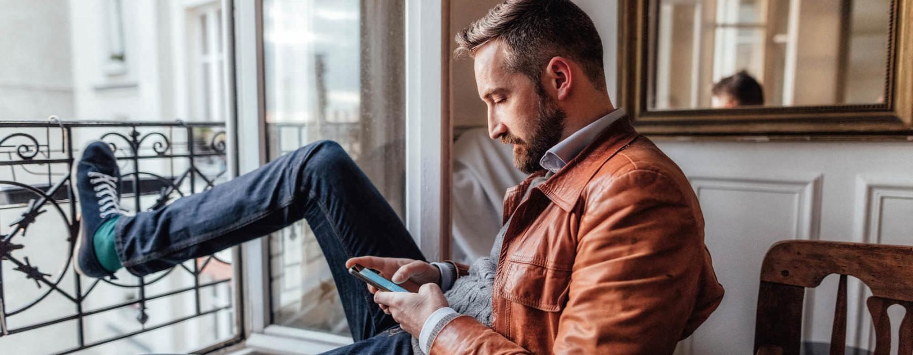 a man sits on a chair and rests his foot on a balcony railing as he looks at his phone in his room